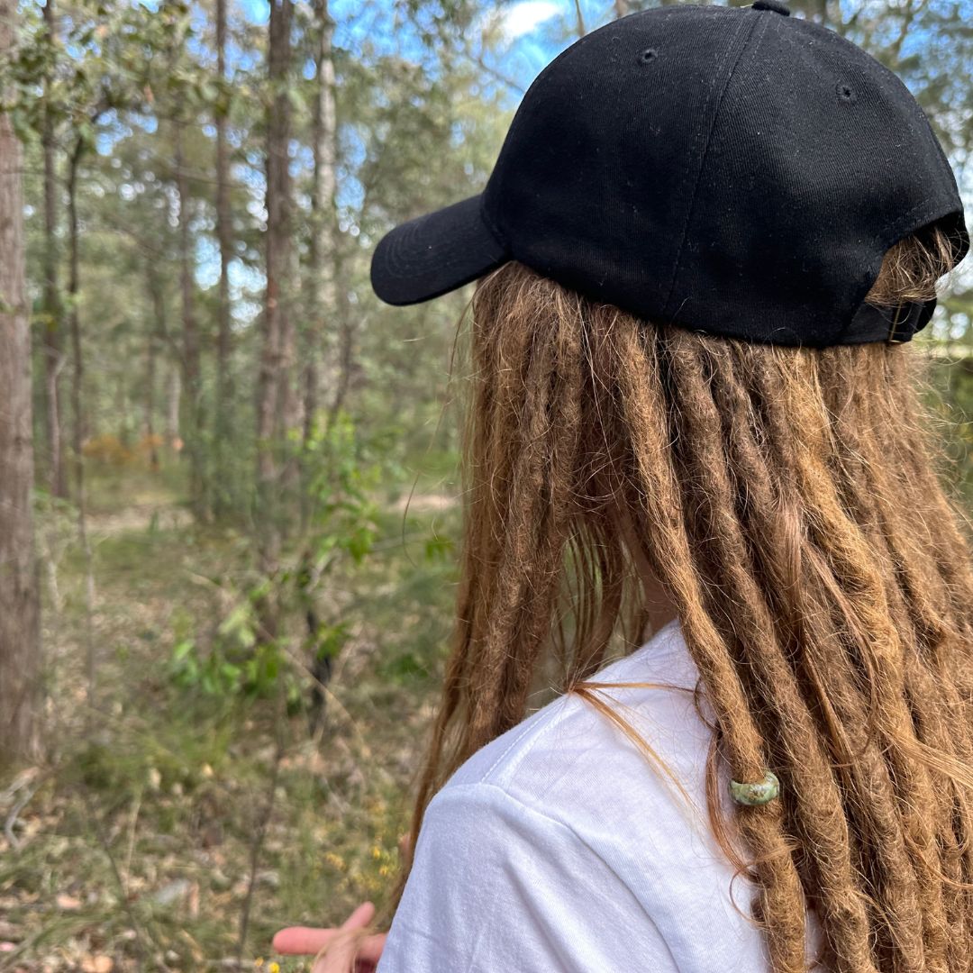 Person with dreadlocks wearing a black cap in a forest setting