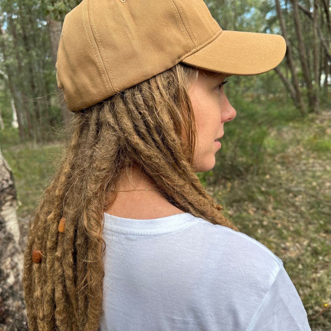 Person with dreadlocks wearing a tan cap in a forest setting