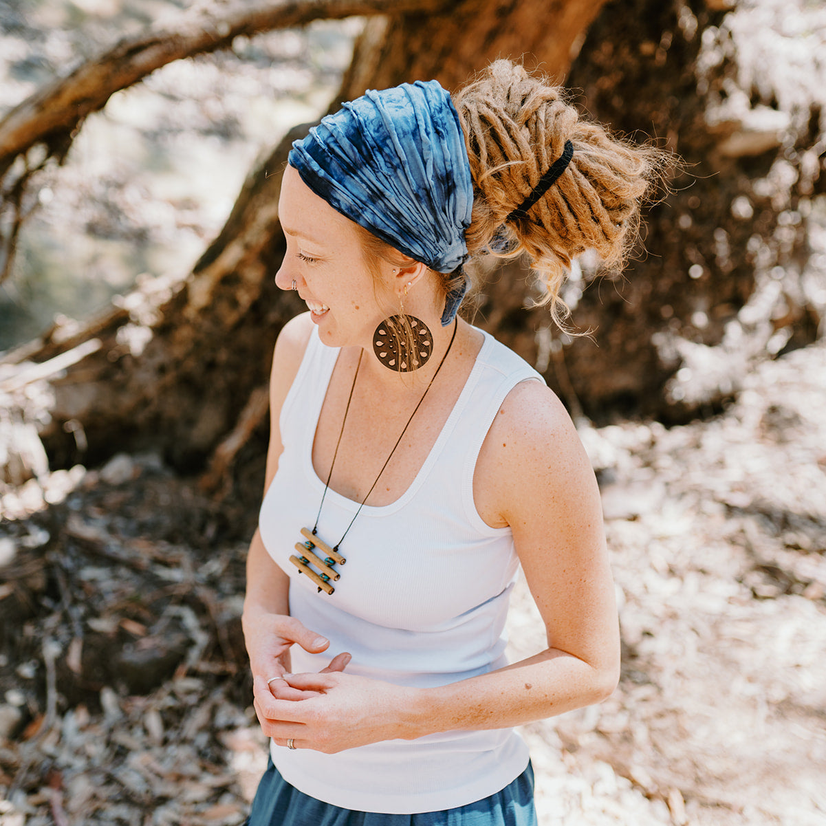Woman with a blue headscarf and white tank top standing in a natural setting.