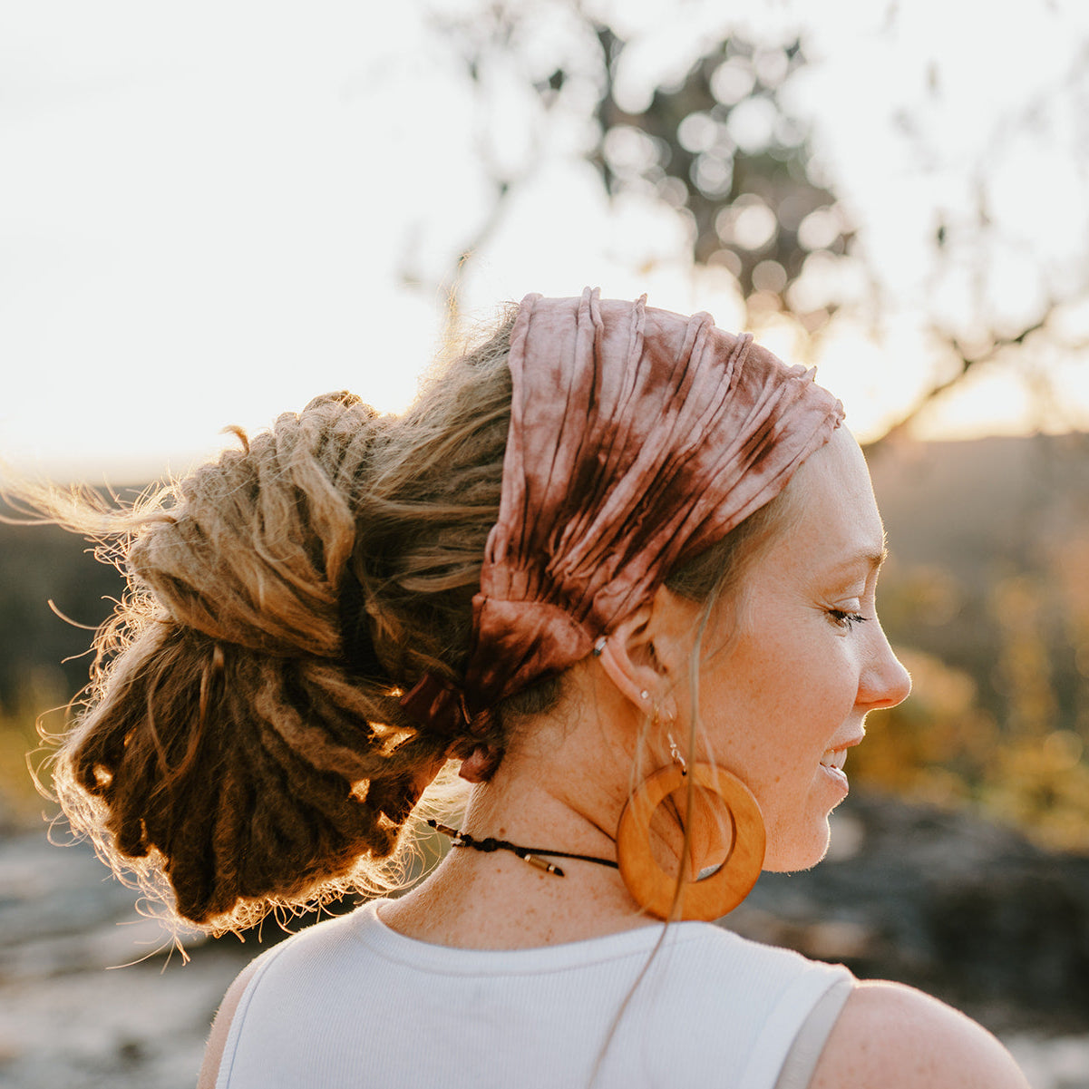 Woman with dreads, hair tie and large earrings standing outdoors with a blurred natural background