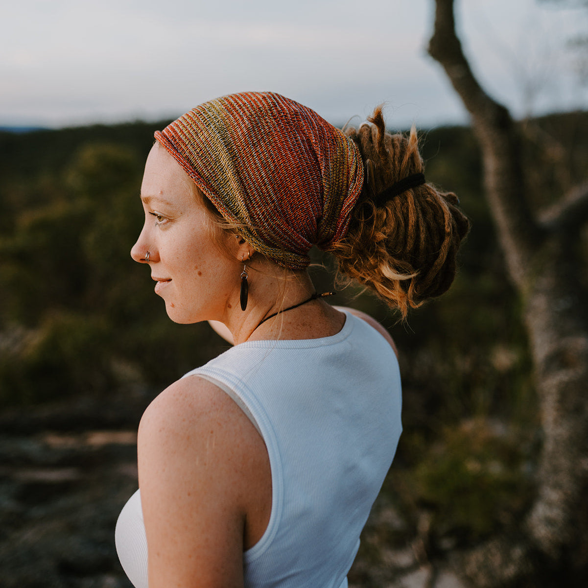 Woman with a colorful headscarf and light blue sleeveless top standing outdoors with trees in the background