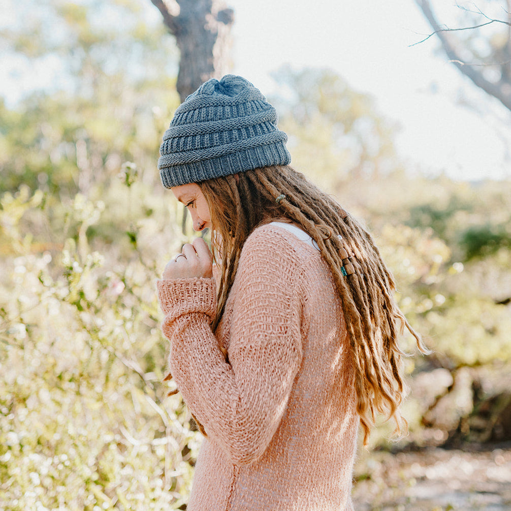 Person with dreadlocks wearing a gray beanie and pink sweater in a natural setting.