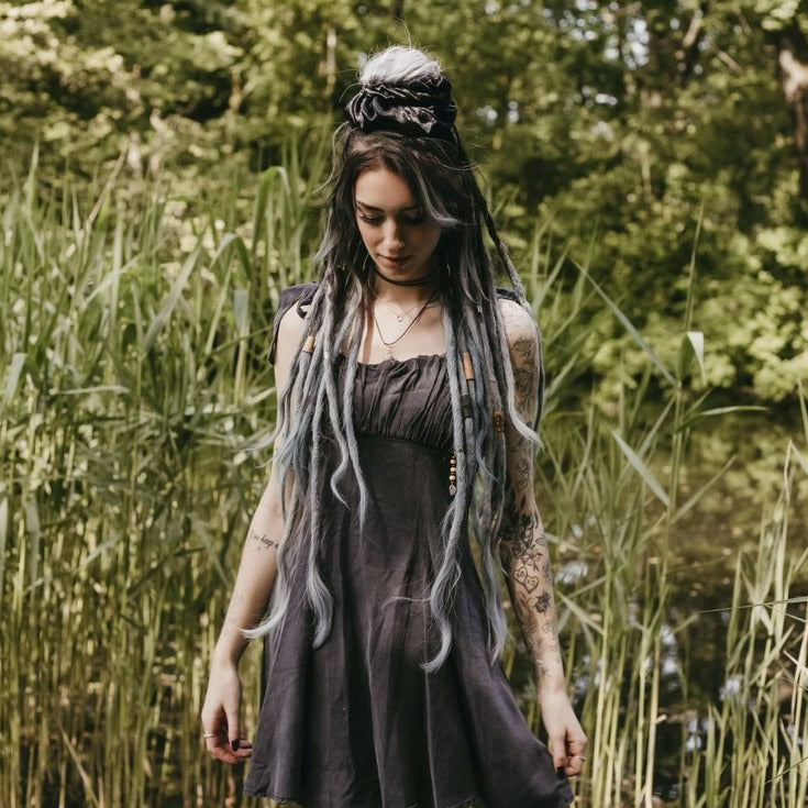 A person with blue dreadlocks wearing a black satin dread tie scrunchie, standing in a natural outdoor setting with greenery in the background.