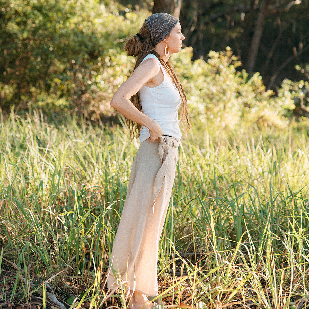 Woman standing in a grassy field wearing a white tank top and beige wrap skirt.